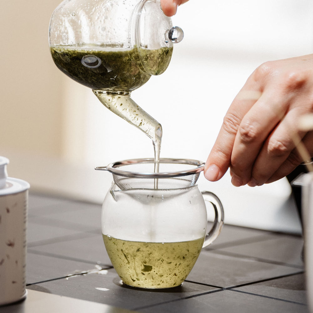 Glasteekanne gießt grünen Kräutertee durch ein Metallsieb in ein Glas auf grauer Fliese neben weißer Teedose.