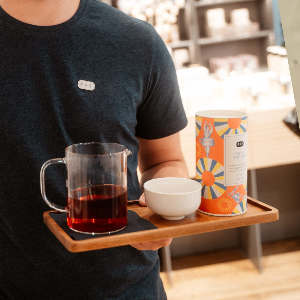 Person im dunkelgrauen P&T-T-Shirt hält Tablett mit Glaskrug, weißer Tasse und 'TOP OF THE DAY'-Dose im Café.