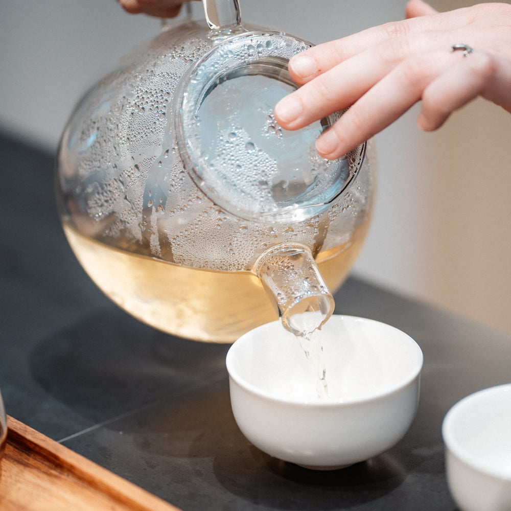 Glasteekanne mit hellem Tee gießt in weiße Keramiktasse auf dunklem Untergrund.