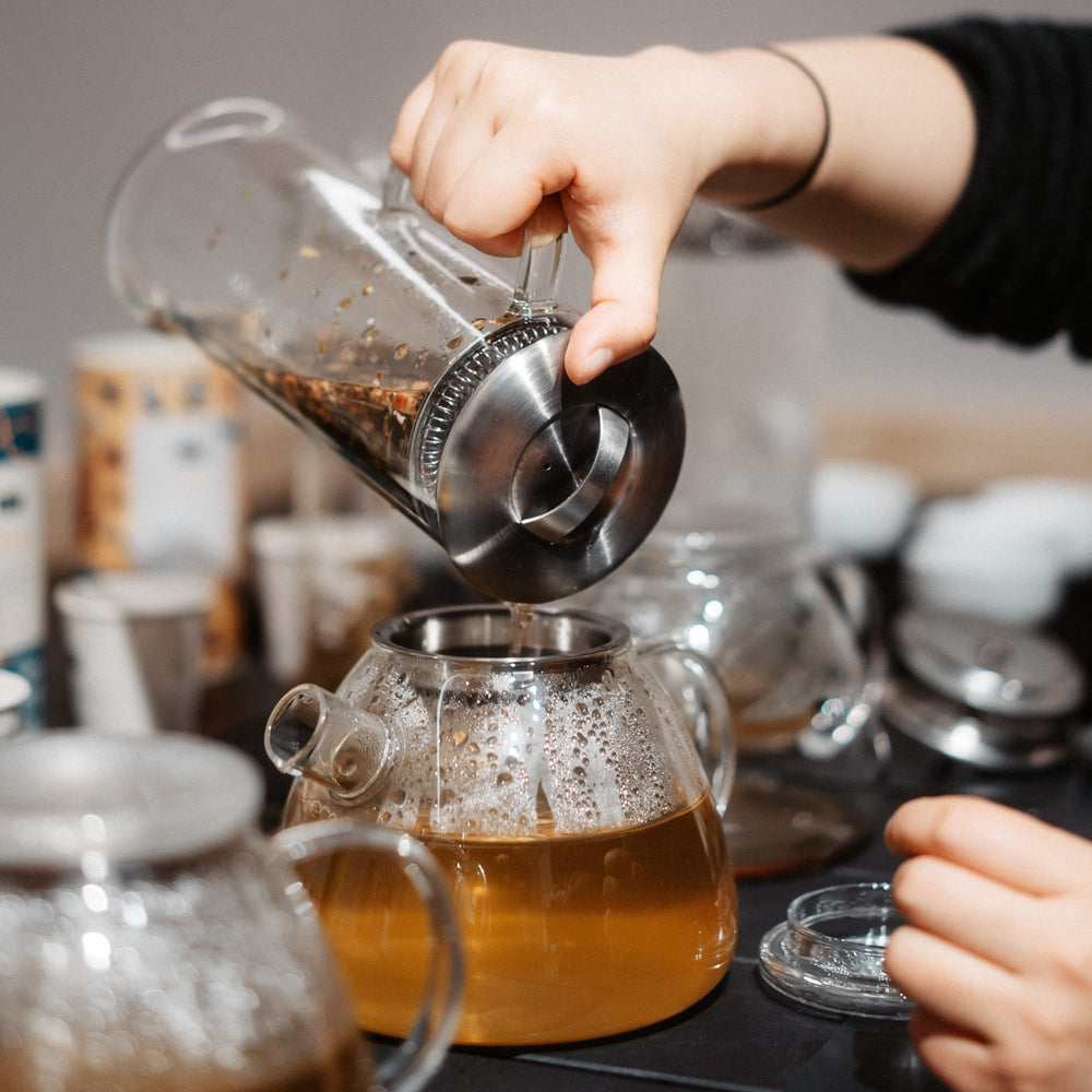 Klare Glasteetasse mit dampfendem Tee, während frischer Tee aus einer Glaskanne mit Metalldeckel eingeschenkt wird.