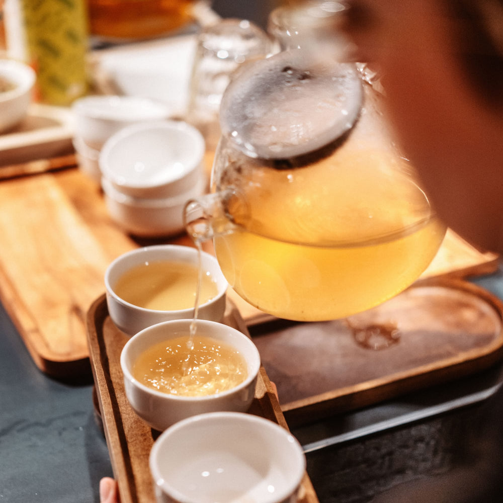 Glasteekanne gießt bernsteinfarbenen Tee in weiße Keramiktasse auf Tablett mit zwei weiteren Tassen.