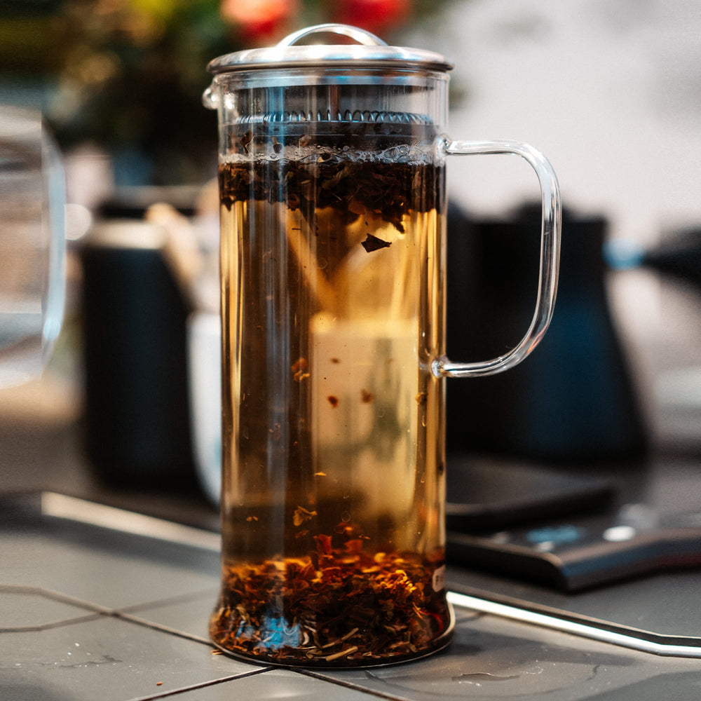 Transparente French Press aus Glas mit Teeblättern und glänzendem Metalldeckel im Vordergrund.