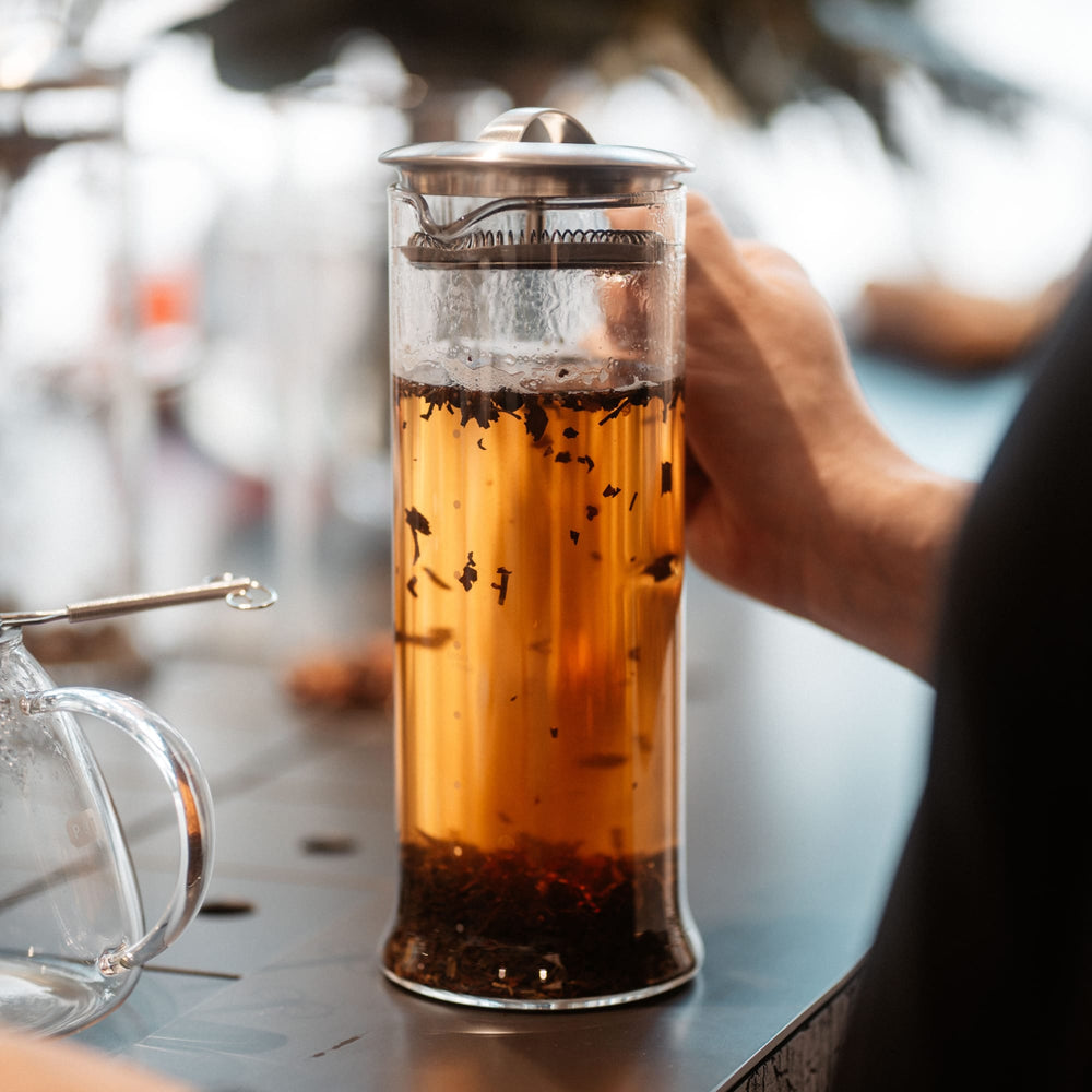 Hohe Glaskanne mit bernsteinfarbenem Tee und Teeblättern in einer French Press neben Glas-Teekännchen.