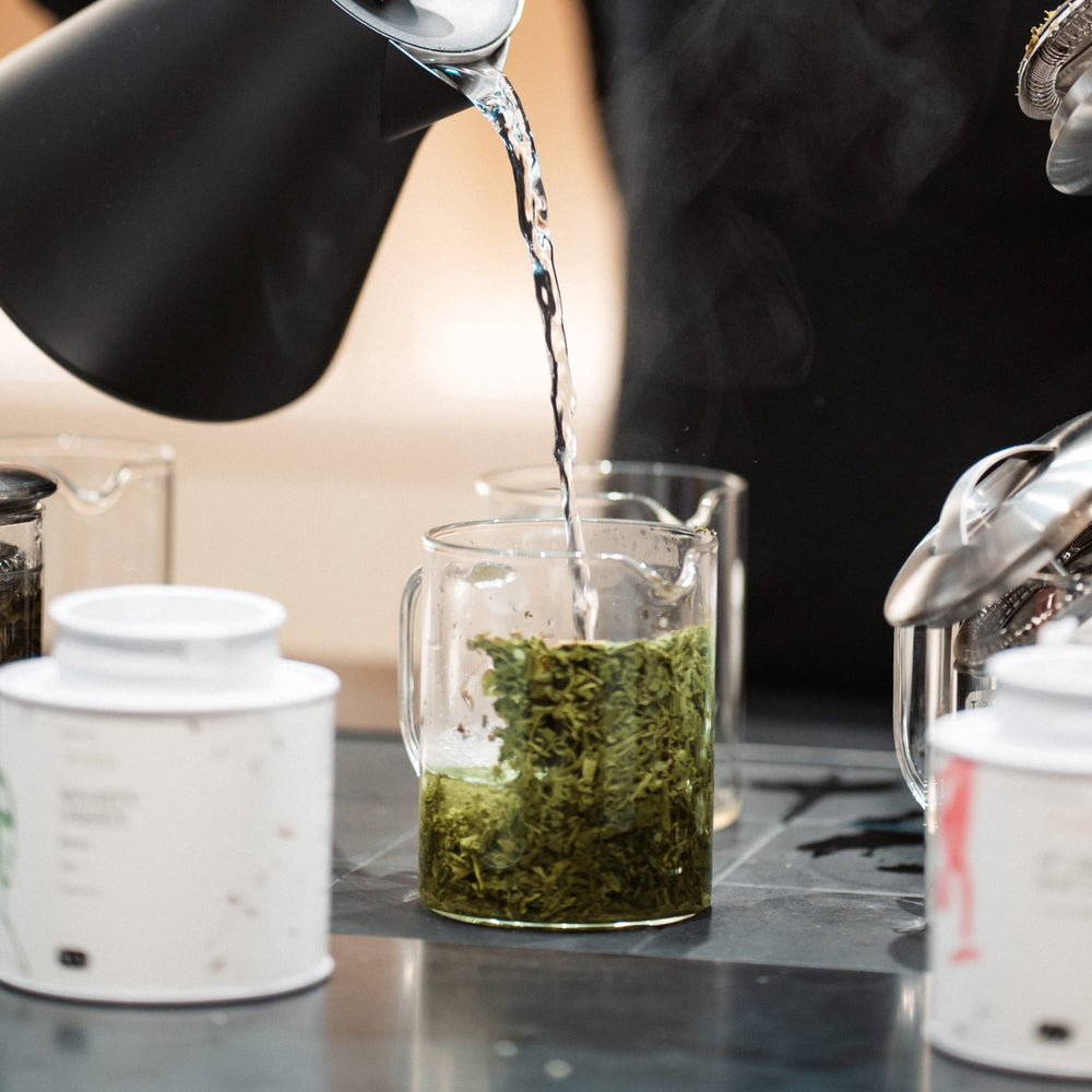 Transparente Glastasse mit grünem Tee, heißes Wasser wird aus schwarzem Wasserkocher eingeschenkt.